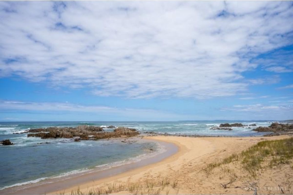 A scenic coastal view featuring a sandy beach, rocky outcrops, and the ocean under a blue sky with scattered clouds.