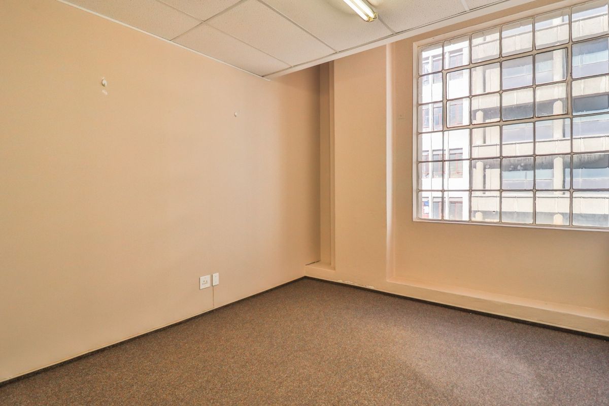 A vacant office space featuring carpeted flooring, a suspended ceiling with integrated lighting, and a large glass block window providing natural light.