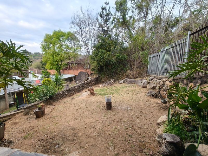 A sloped backyard garden featuring a stone retaining wall, metal security fencing, and established trees, offering potential for landscaping and outdoor improvement.