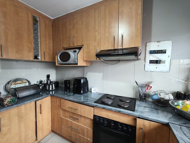 A functional kitchen featuring wood-finish cabinetry, stone-look laminate countertops, integrated appliances, and a tiled backsplash with an exposed electrical distribution board.