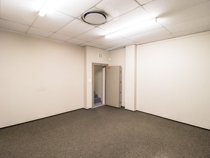 A clean, empty utility or storage room featuring a heavy-duty security door, suspended ceiling with fluorescent lighting, and carpeted flooring.