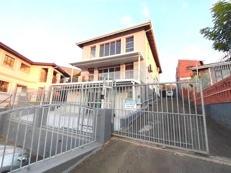 Front view of a multi-story residential building featuring a metal security gate, concrete driveway, and a balcony with sliding glass doors.