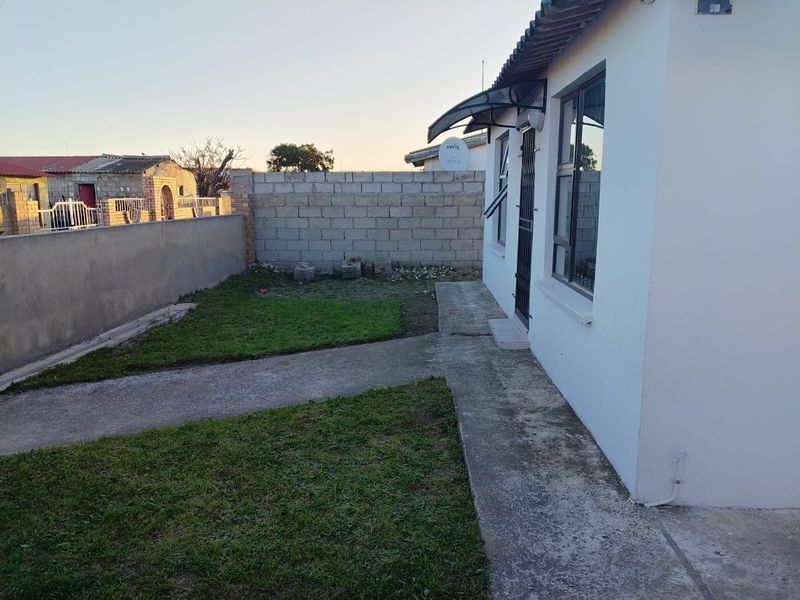 A rear exterior view of the property featuring a concrete walkway, a small lawn area, and a perimeter boundary wall.