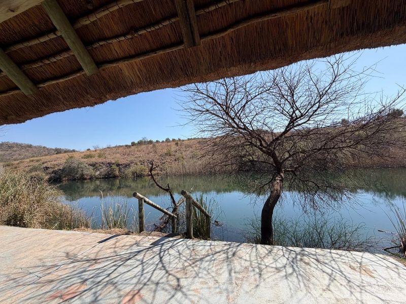 A covered patio area with a traditional thatch roof, featuring a concrete floor and timber railing overlooking a tranquil farm dam.
