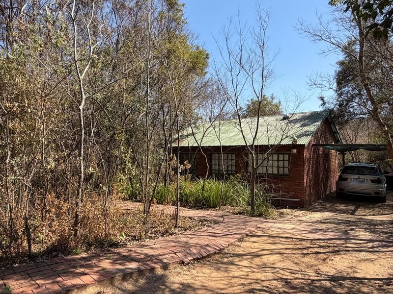A rustic brick farmhouse exterior featuring a corrugated metal roof, grid-pattern windows, and a paved walkway surrounded by mature trees and natural landscaping.