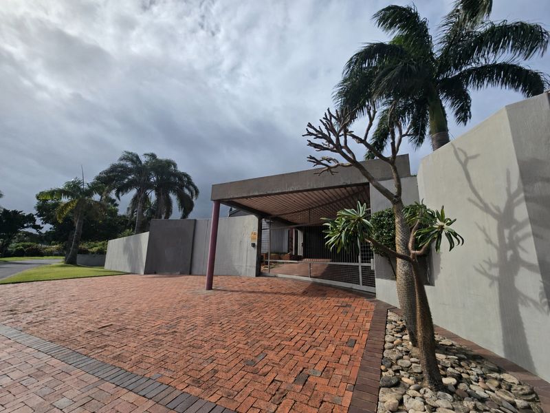 Front exterior view of the property featuring a brick driveway, covered entrance area, and concrete perimeter walls with established landscaping.