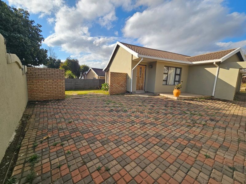Front exterior view of a single-story house featuring a paved driveway, brick boundary walls, and a tiled roof with security gate access.