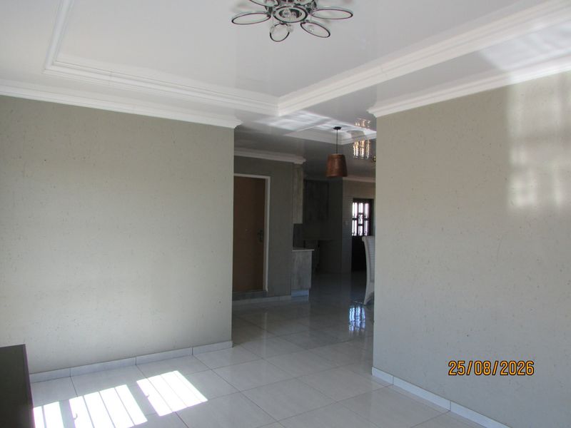 A bright, open-plan living area featuring neutral wall tones, polished tiled flooring, and decorative coved ceilings with modern lighting.