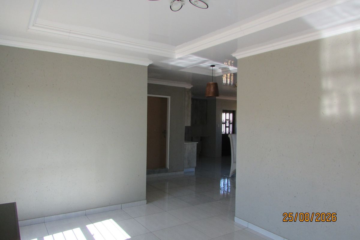 A bright, open-plan living area featuring neutral wall tones, polished tiled flooring, and decorative coved ceilings with modern lighting.