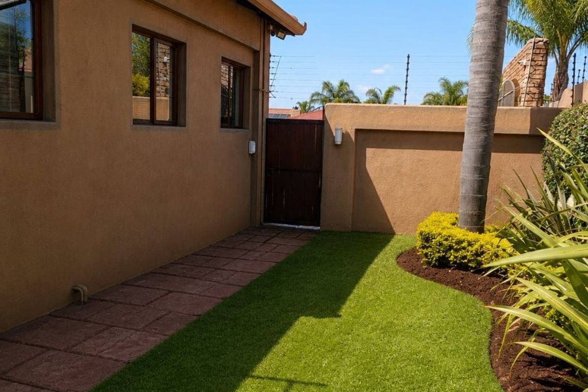 A well-maintained rear garden area featuring a paved walkway, artificial lawn, and secure boundary wall with a wooden side gate.