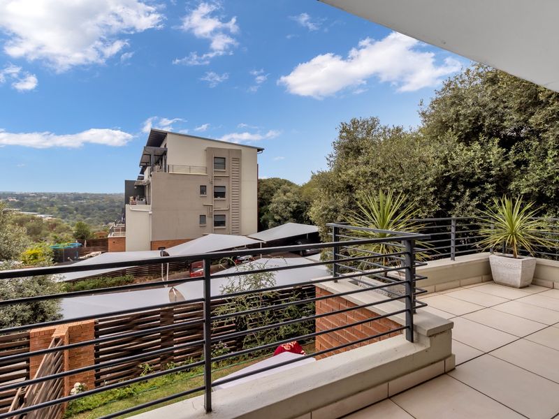 A spacious covered balcony featuring tiled flooring and a metal balustrade, offering elevated views of the surrounding complex and landscape.