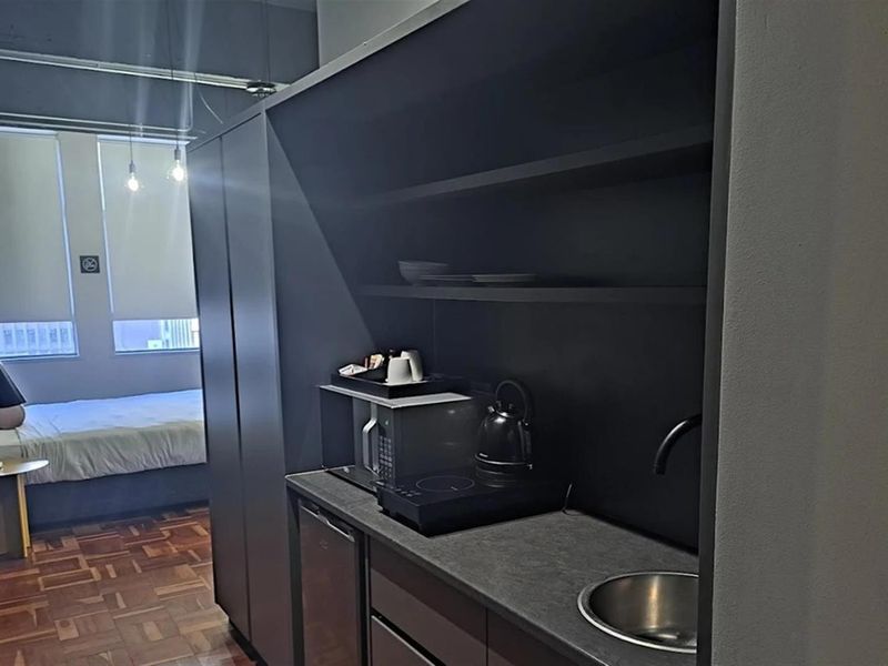 Compact kitchenette featuring dark cabinetry, integrated sink, and countertop, set within an open-plan apartment with parquet flooring.