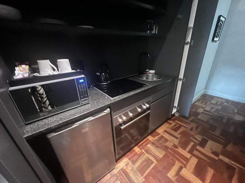 Compact kitchenette featuring a built-in oven, electric hob, sink, and stone countertop, complemented by dark cabinetry and classic parquet flooring.