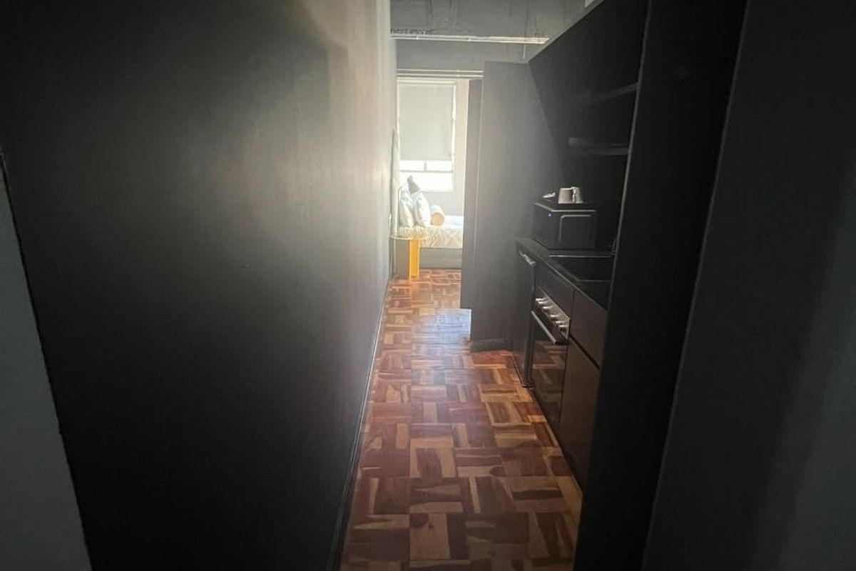 A narrow galley-style kitchen featuring dark cabinetry, built-in oven, and classic parquet flooring, leading towards a bedroom area.