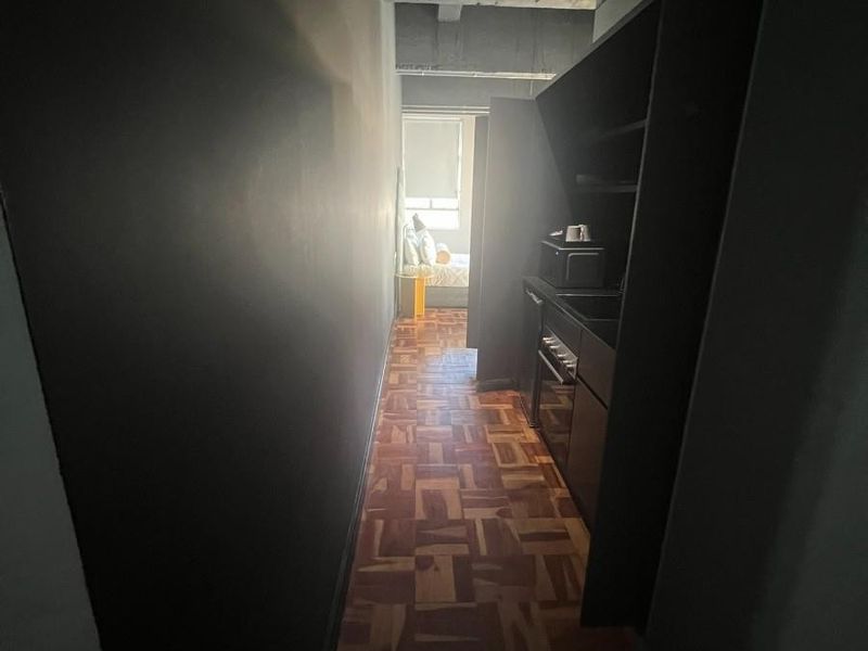 A narrow galley-style kitchen featuring dark cabinetry, built-in oven, and classic parquet flooring, leading towards a bedroom area.