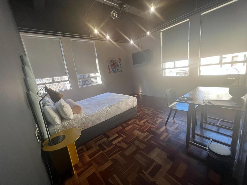 Spacious bedroom featuring classic parquet flooring, high ceilings with industrial-style pendant lighting, and large windows fitted with roller blinds for natural light control.