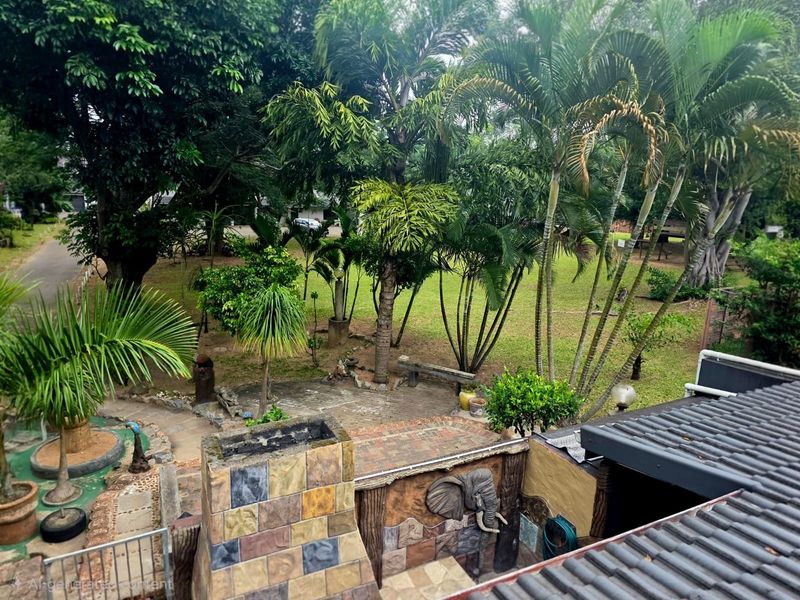 An elevated view of a rear garden featuring a stone-clad outdoor fireplace, tiled patio area, and mature landscaping.