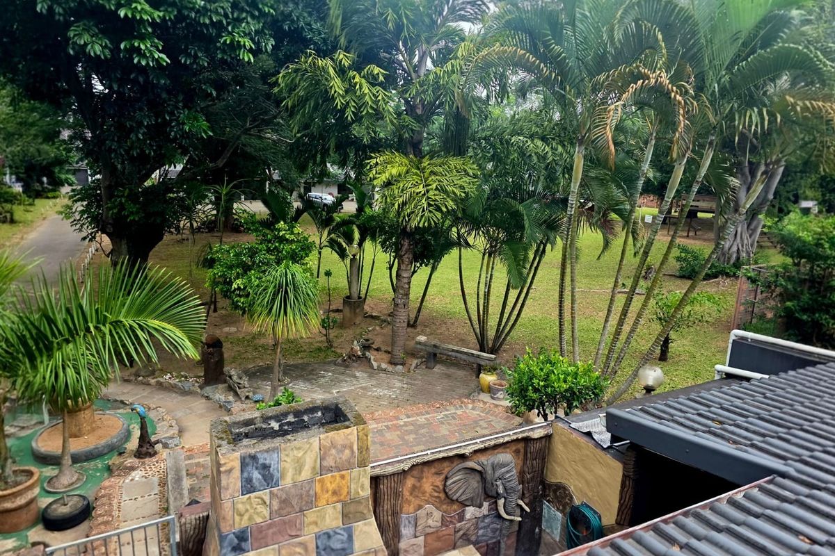 An elevated view of a rear garden featuring a stone-clad outdoor fireplace, tiled patio area, and mature landscaping.