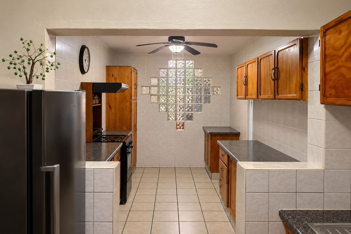 A functional kitchen featuring wooden cabinetry, tiled surfaces, a unique glass block window feature, and a ceiling fan.