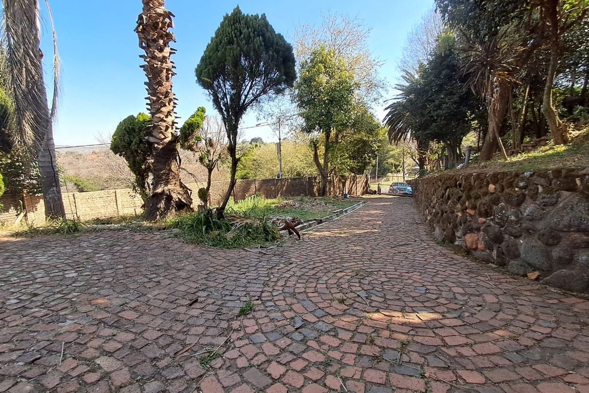 A spacious rear garden area featuring a cobblestone driveway, stone retaining walls, and established mature trees.