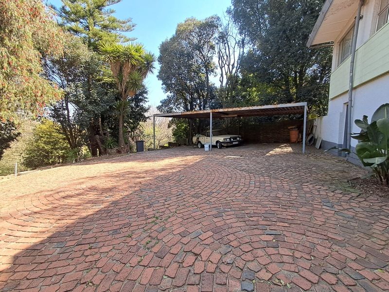 Spacious brick-paved driveway featuring a covered carport area, surrounded by mature trees and established garden landscaping.