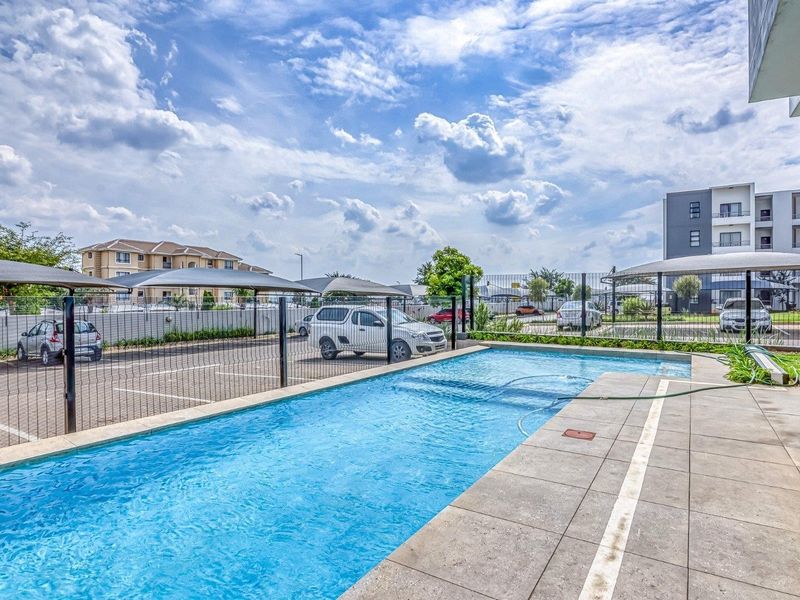 A well-maintained swimming pool area featuring a tiled deck, perimeter fencing, and adjacent covered parking facilities within the apartment complex.
