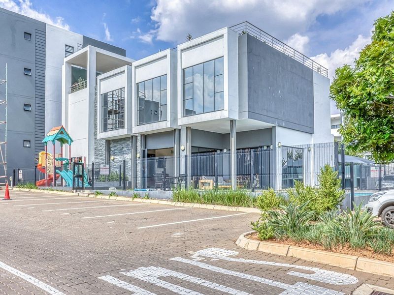 A modern apartment building exterior featuring contemporary architectural design, large glass windows, and a paved parking area with perimeter fencing.