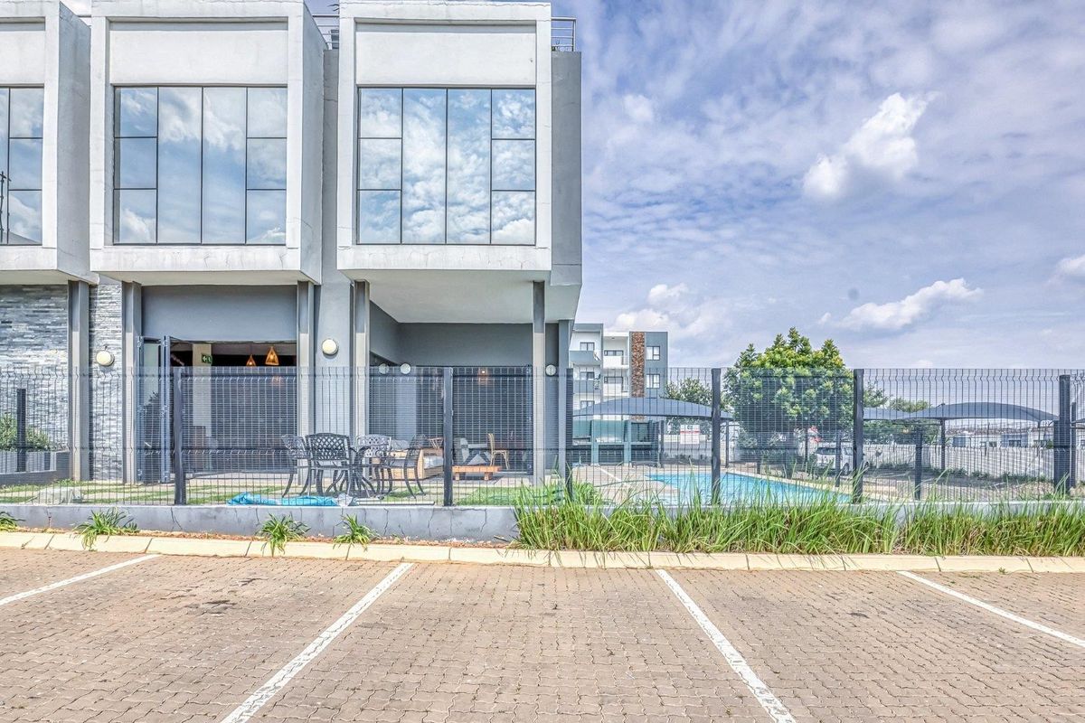 Rear view of a modern apartment complex featuring a paved parking area, wire mesh fencing, and a communal swimming pool.
