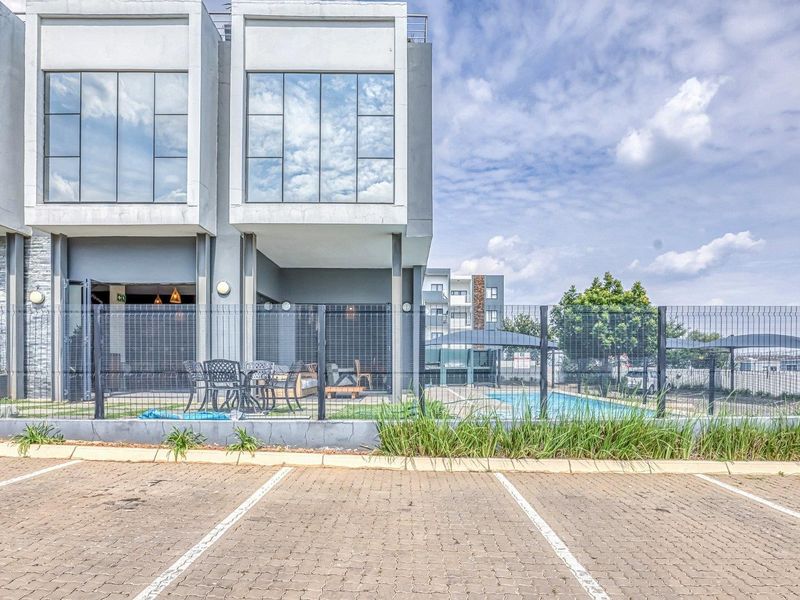 Rear view of a modern apartment complex featuring a paved parking area, wire mesh fencing, and a communal swimming pool.