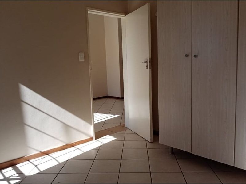 A bright, empty bedroom featuring neutral tiled flooring and a built-in wardrobe, with a doorway leading to an adjacent hallway.
