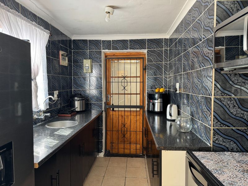 A compact kitchen featuring dark cabinetry, granite-look laminate countertops, and distinctive patterned wall tiles, leading to an exterior door with a security gate.