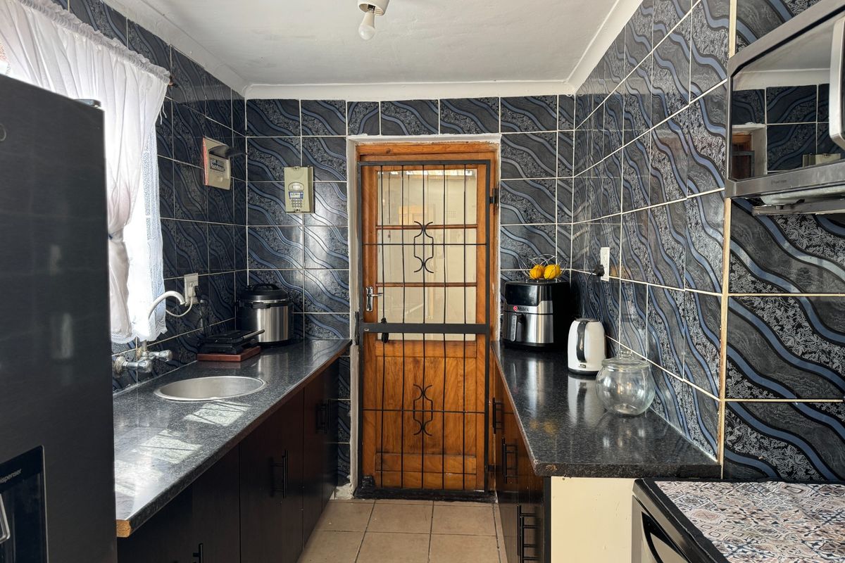 A compact kitchen featuring dark cabinetry, granite-look laminate countertops, and distinctive patterned wall tiles, leading to an exterior door with a security gate.