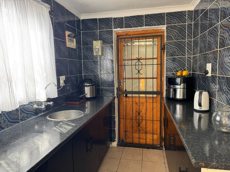 A functional scullery featuring dark cabinetry, granite countertops, stainless steel sink, and full-height decorative wall tiling with a secure wooden door.
