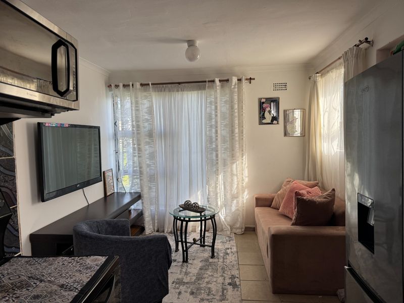 Compact open-plan living area featuring tiled flooring, neutral walls, and natural light through large window treatments.