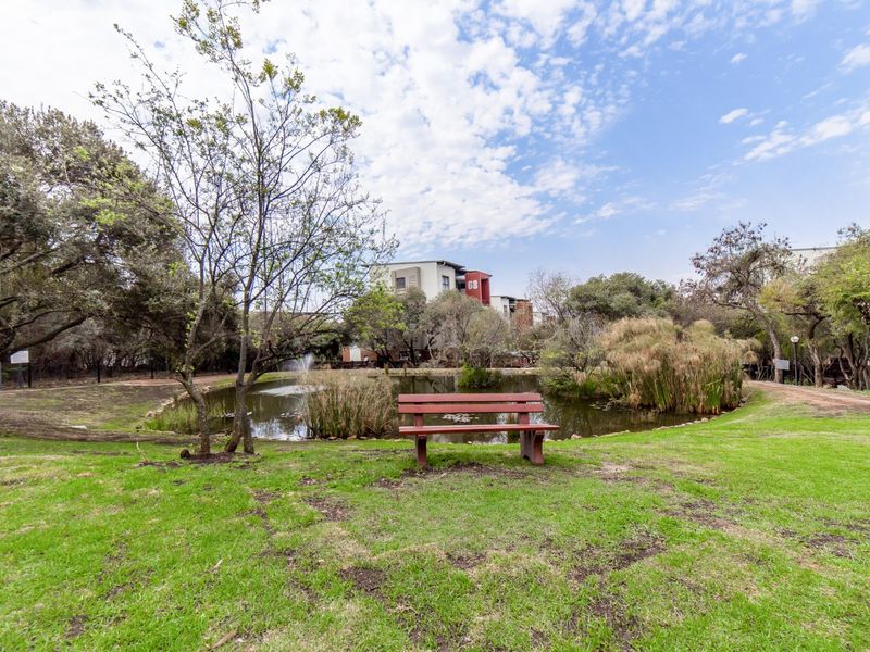 A peaceful communal garden area featuring a landscaped lawn, a pond with a water feature, and mature trees within the apartment complex grounds.