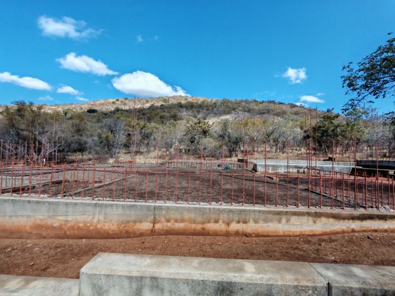 A construction site showing an exposed concrete foundation and steel reinforcement bars set against a natural hillside backdrop.