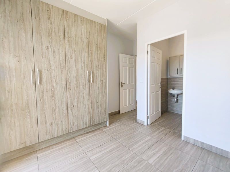 A clean, empty bedroom featuring modern wood-look built-in cupboards, neutral tiled flooring, and direct access to an en-suite bathroom.