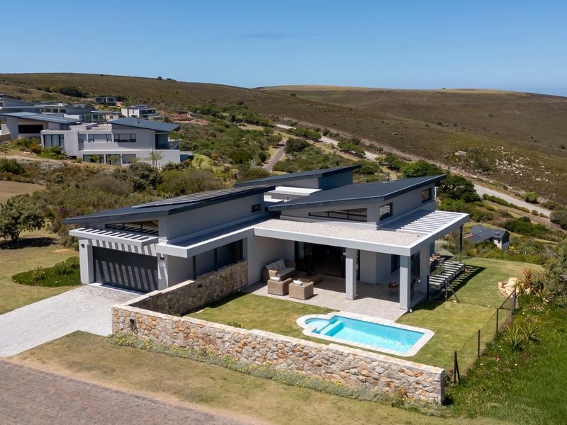 Aerial view of a modern contemporary home featuring a swimming pool, stone wall, covered patio, and solar panels in a scenic estate setting.