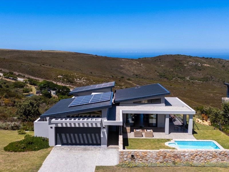 Aerial view of a modern home featuring a metal roof with solar panels, a swimming pool, stone retaining wall, and a paved driveway.