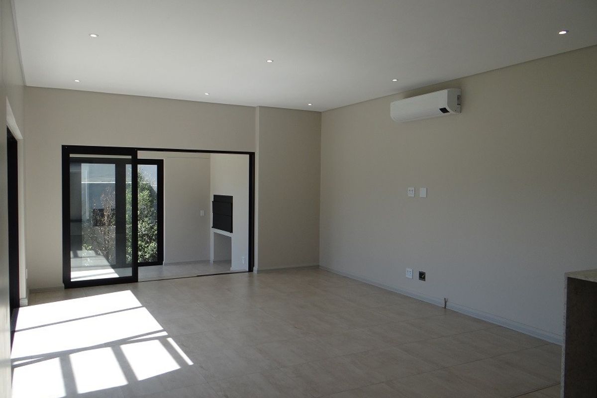 A spacious, modern, and empty living area featuring neutral tiled floors, recessed lighting, and a wall-mounted air conditioning unit with access to an outdoor space.