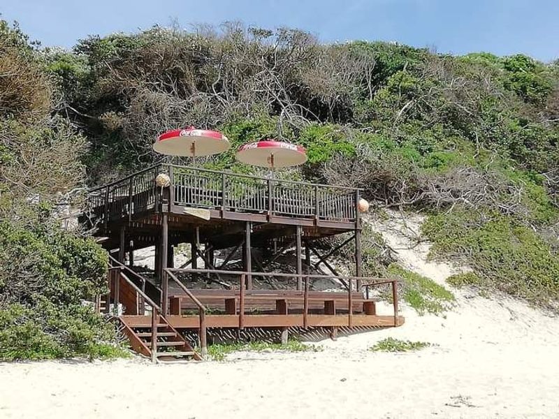 An elevated timber deck structure with built-in bench seating, situated on a sandy coastal dune area.