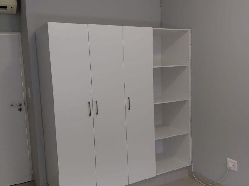 A clean bedroom featuring a modern white built-in wardrobe with integrated open shelving, tiled floors, and a wall-mounted air conditioning unit.