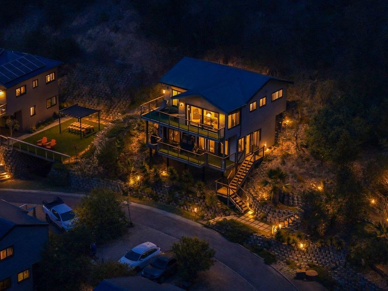 An elevated night-time aerial view of a modern multi-level house featuring extensive decking, integrated outdoor lighting, and terraced landscaping.
