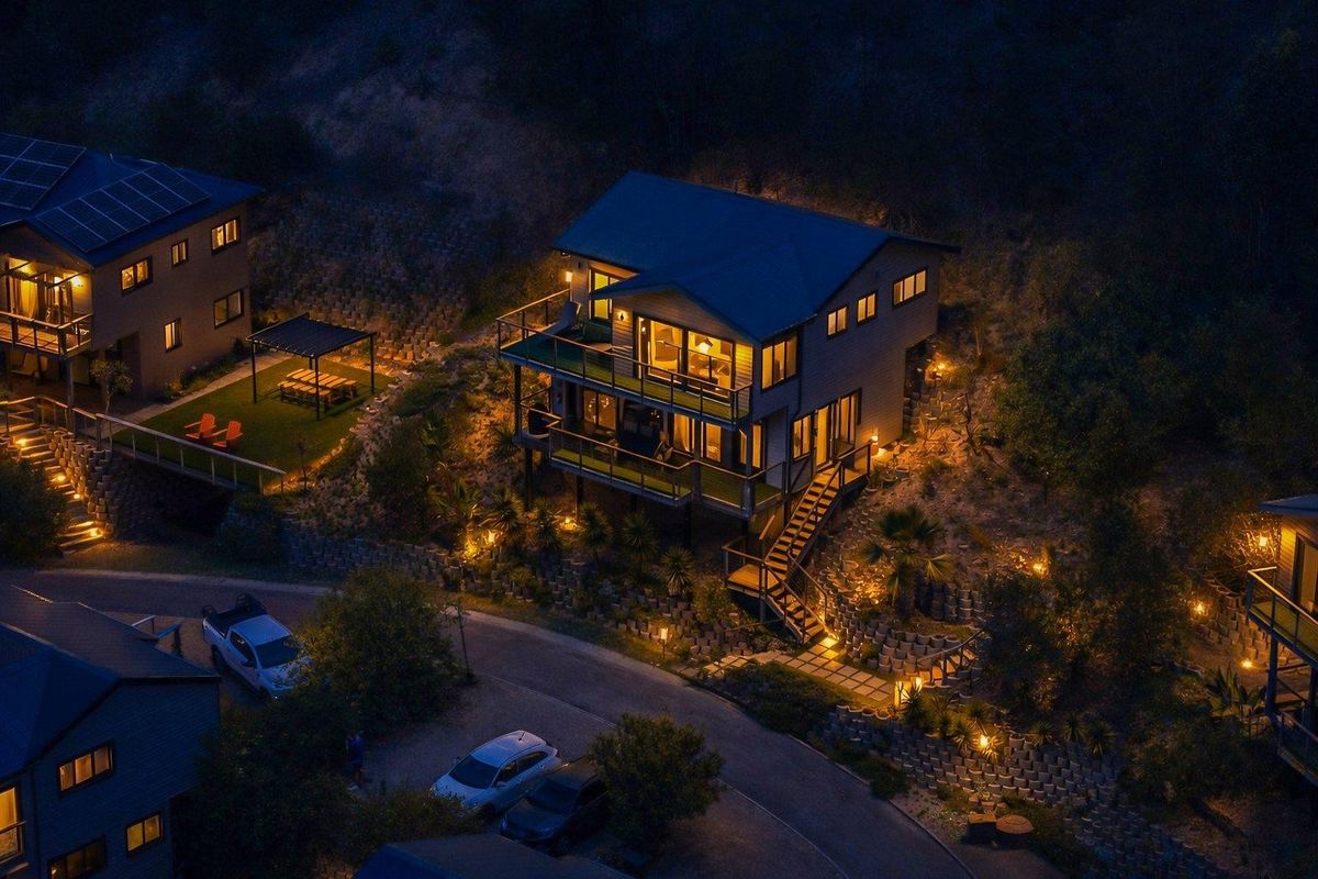 An elevated night-time aerial view of a modern multi-level house featuring extensive decking, integrated outdoor lighting, and terraced landscaping.