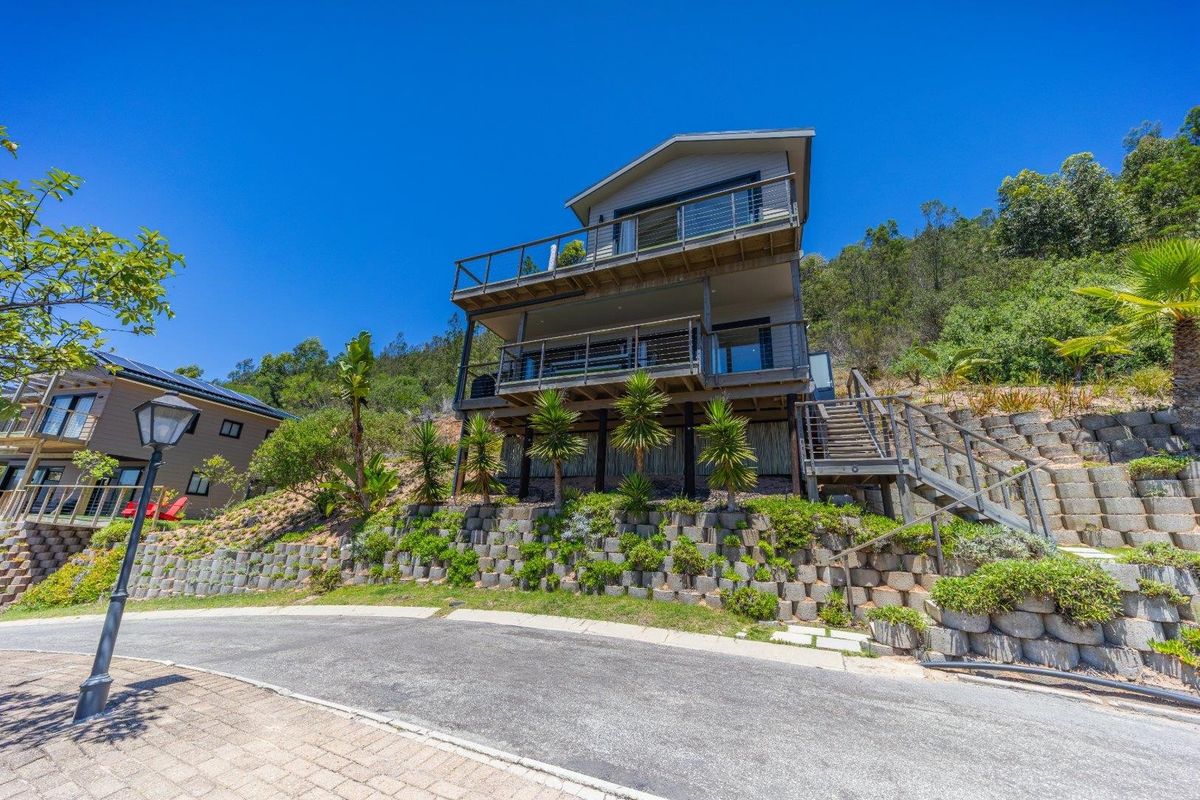 Rear elevation of a multi-level house featuring timber decking, steel balustrades, and a tiered retaining wall set against a hillside.