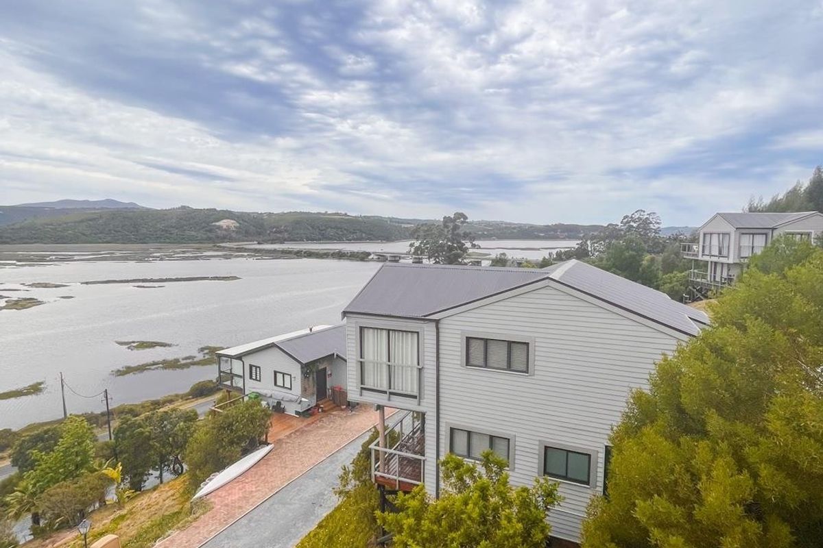 Aerial view of a modern weatherboard house featuring a metal roof, paved driveway, and balcony, set against a scenic estuary landscape.