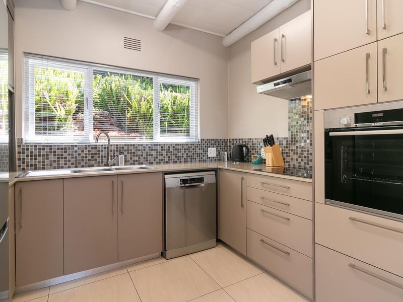 Modern kitchen featuring neutral cabinetry, mosaic tile backsplash, integrated stainless steel appliances, and tiled flooring with natural light from a garden-facing window.