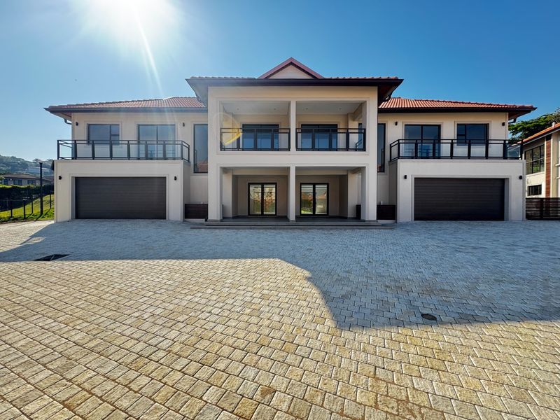 Front elevation of a modern townhouse complex featuring a large paved driveway, double garages, and upper-level balconies with metal railings.