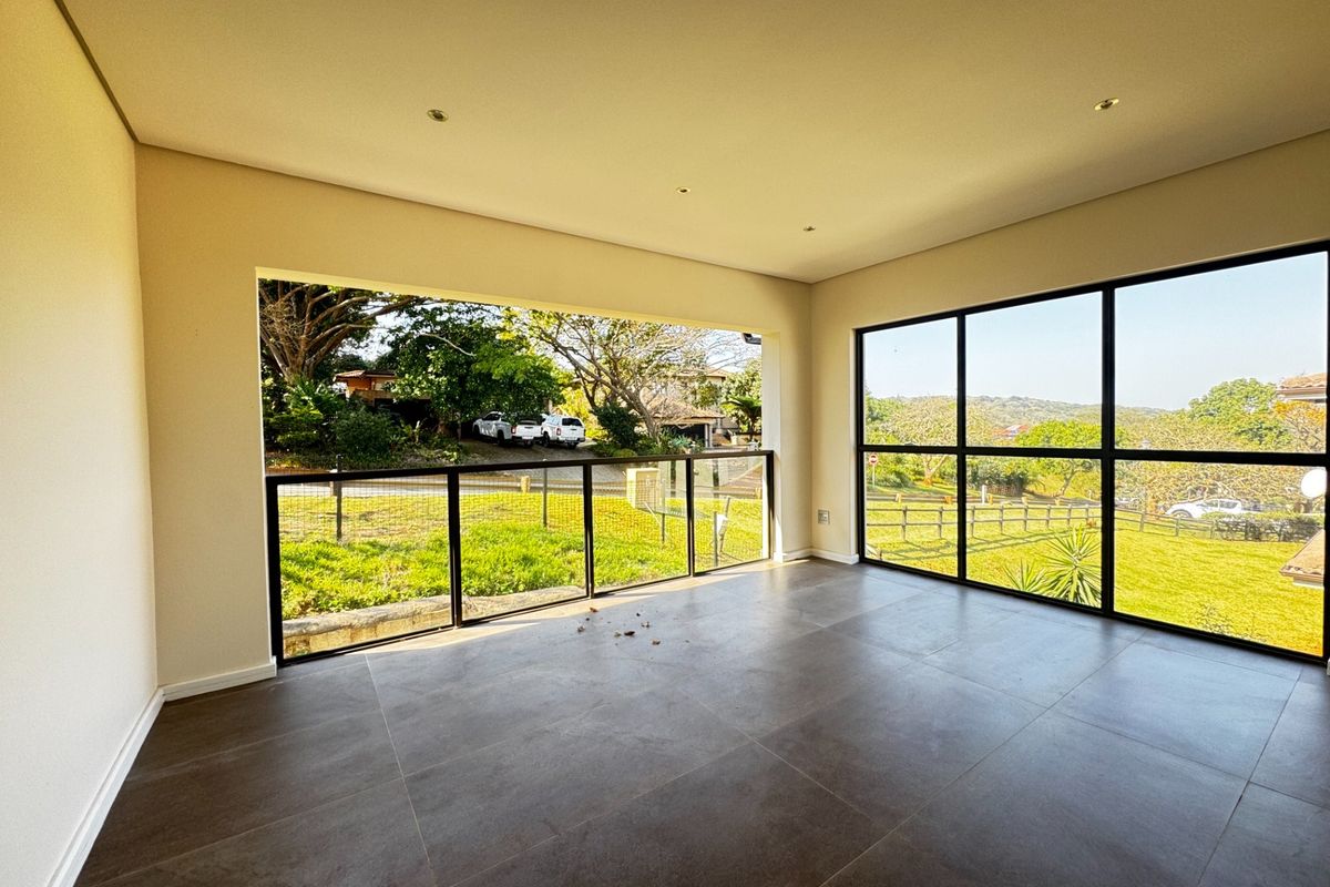 A bright, empty living area featuring large format floor tiles, recessed lighting, and expansive black-framed windows offering scenic garden views.