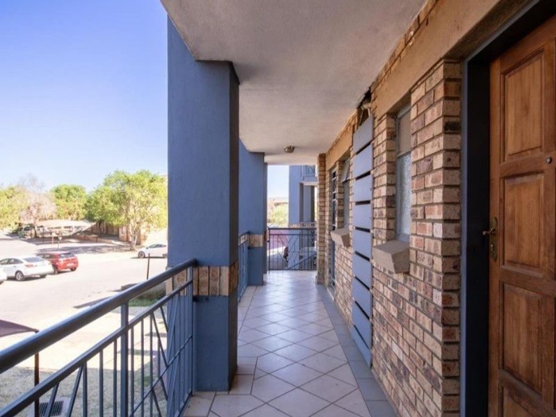 A covered balcony featuring tiled flooring, a metal balustrade, and a timber entrance door, overlooking the apartment complex parking area.